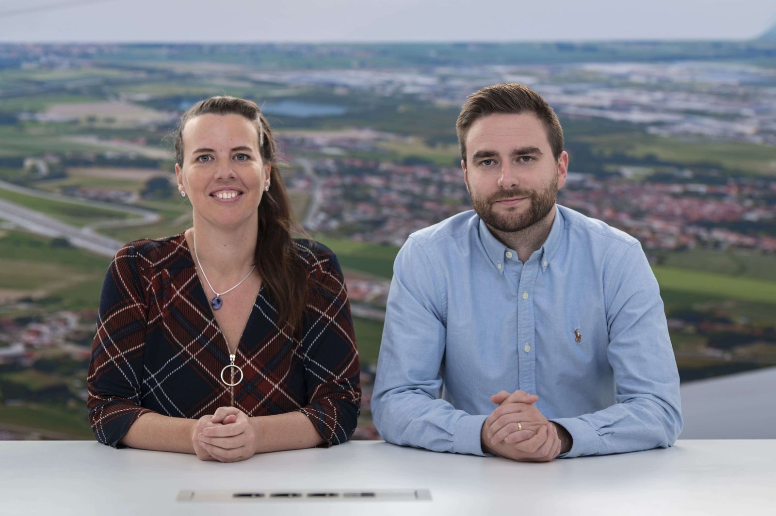 Een vrouw en een man zitten naast elkaar aan een wit bureau en poseren voor een foto, met op de achtergrond een wazig luchtfoto van een landschap.