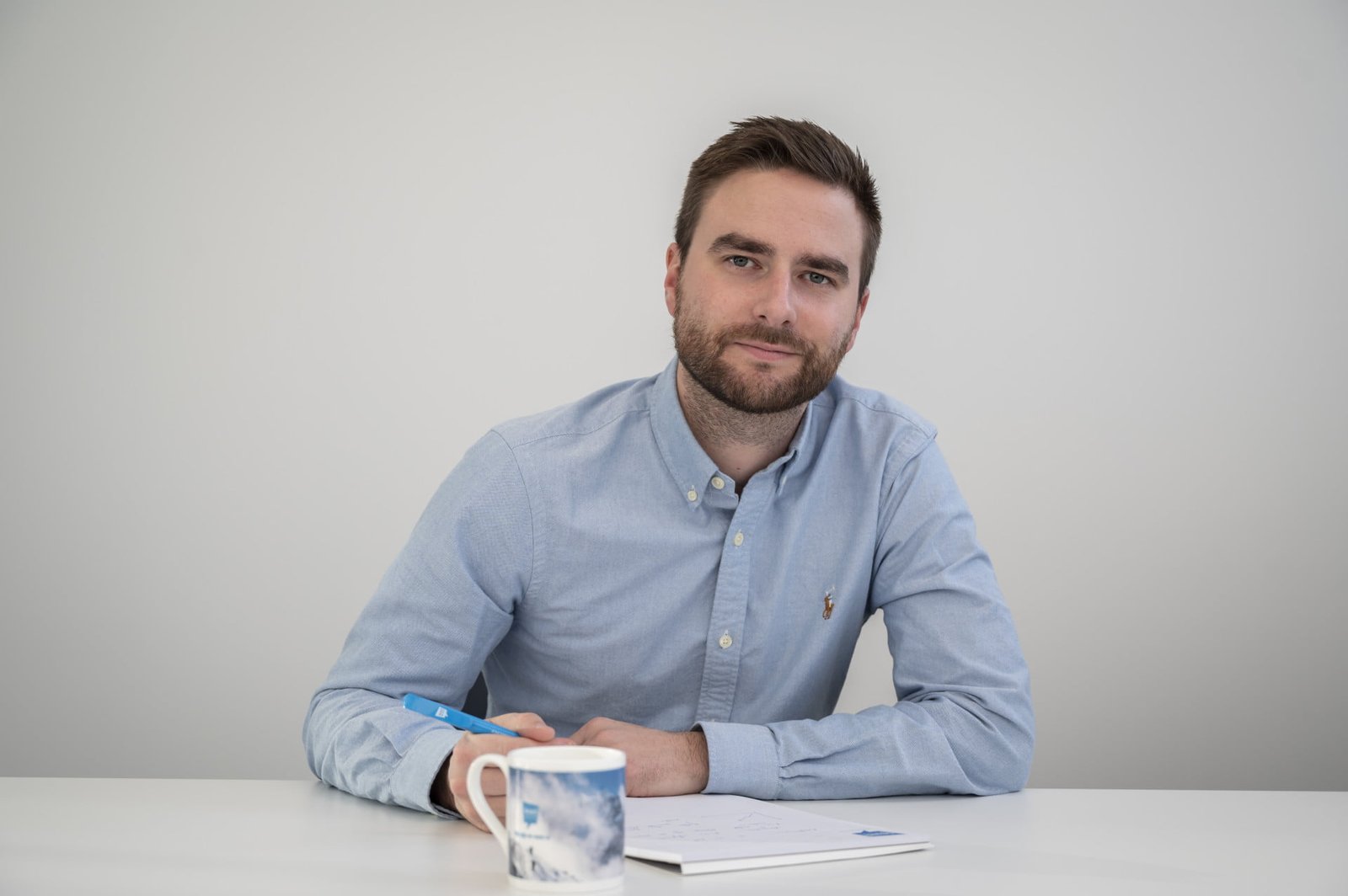 Een man met een baard en een blauw shirt zit achter een bureau, met een pen in zijn hand. Voor hem ligt een notitieboekje en er staat een mok in de buurt.