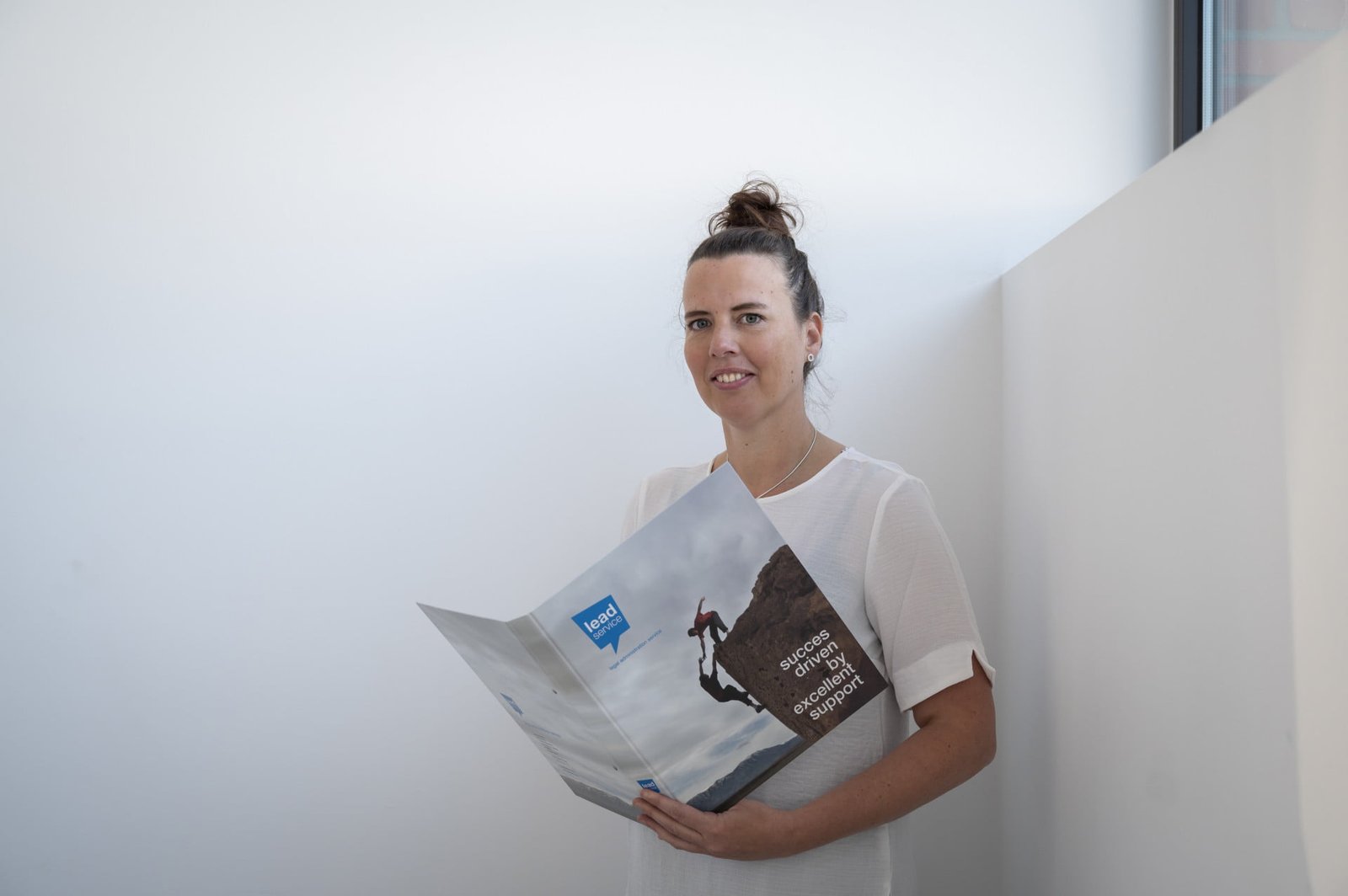 Een persoon in een wit overhemd staat bij een muur en houdt een opengeslagen brochure vast met een afbeelding van een bergbeklimmer op de omslag.